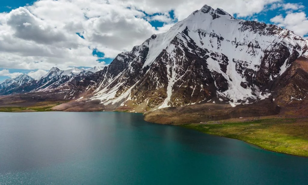 Karambar Lake in the foreground and a snow capped peak in the background with mostly cloudy sky captured from a drone