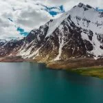 Karambar Lake in the foreground and a snow capped peak in the background with mostly cloudy sky captured from a drone