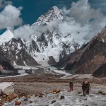 A group of trekkers and porter walking on icy glacier towards the camera with towering K2 peak in the background partially covered by the clouds making it look dramatic