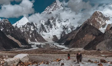 A group of trekkers and porter walking on icy glacier towards the camera with towering K2 peak in the background partially covered by the clouds making it look dramatic