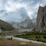 Tall rock granite towers on both sides if the Nangma Valley in the background, cloudy day and green grass in the foreground with 3 locals standing in the distance