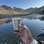 A hand holding a tea cup with landscape of Kharrari in the background inlcuding river and mountains in Balochistan