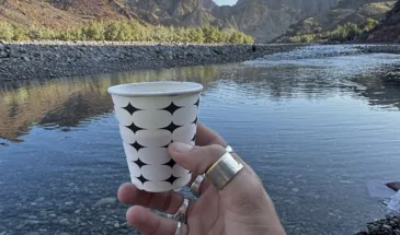 A hand holding a tea cup with landscape of Kharrari in the background inlcuding river and mountains in Balochistan