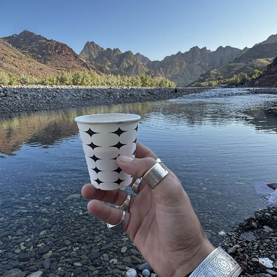 A hand holding a tea cup with landscape of Kharrari in the background inlcuding river and mountains in Balochistan